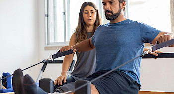 Hombre en ropa deportiva haciendo ejercicios con una mujer que lo observa al lado.